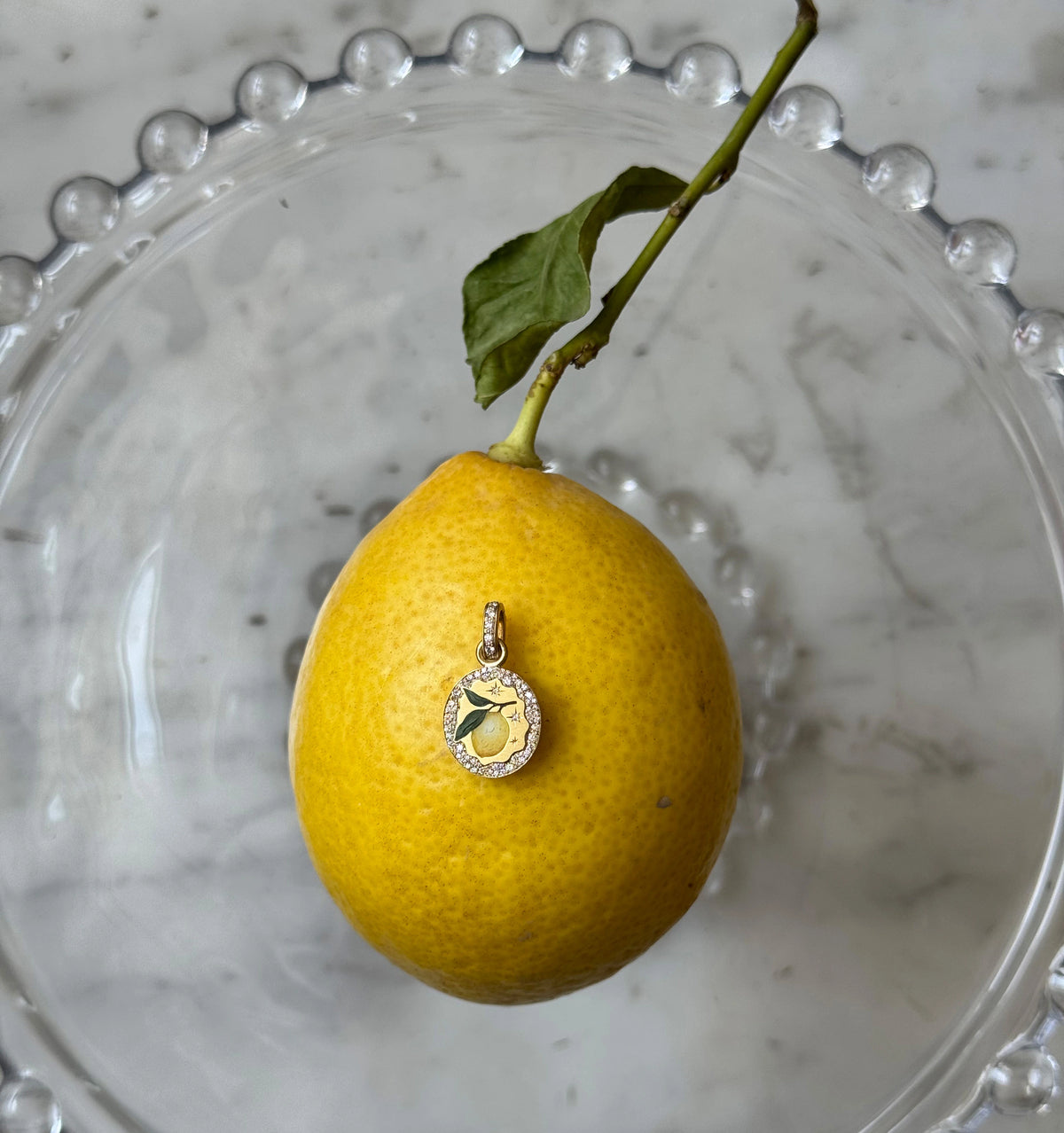 Gold pendant featuring a lemon in enamel on a lemon, set in a glass dish with decorative edges.