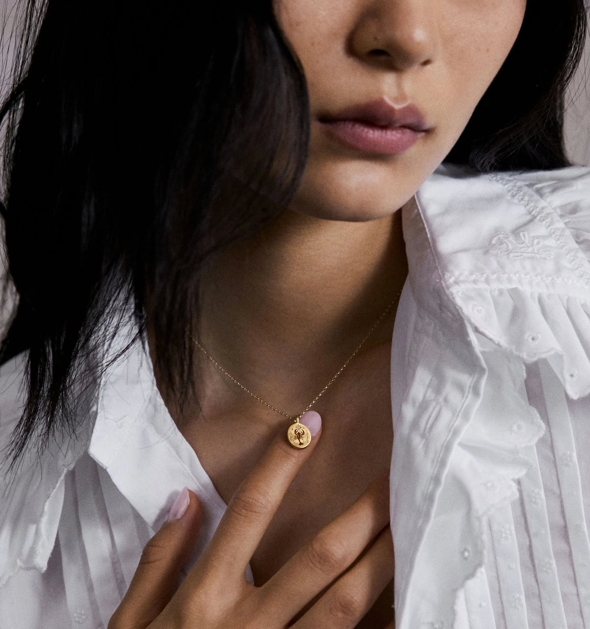 Woman wearing a gold necklace with a circular pendant, touching it gently while dressed in a white shirt.