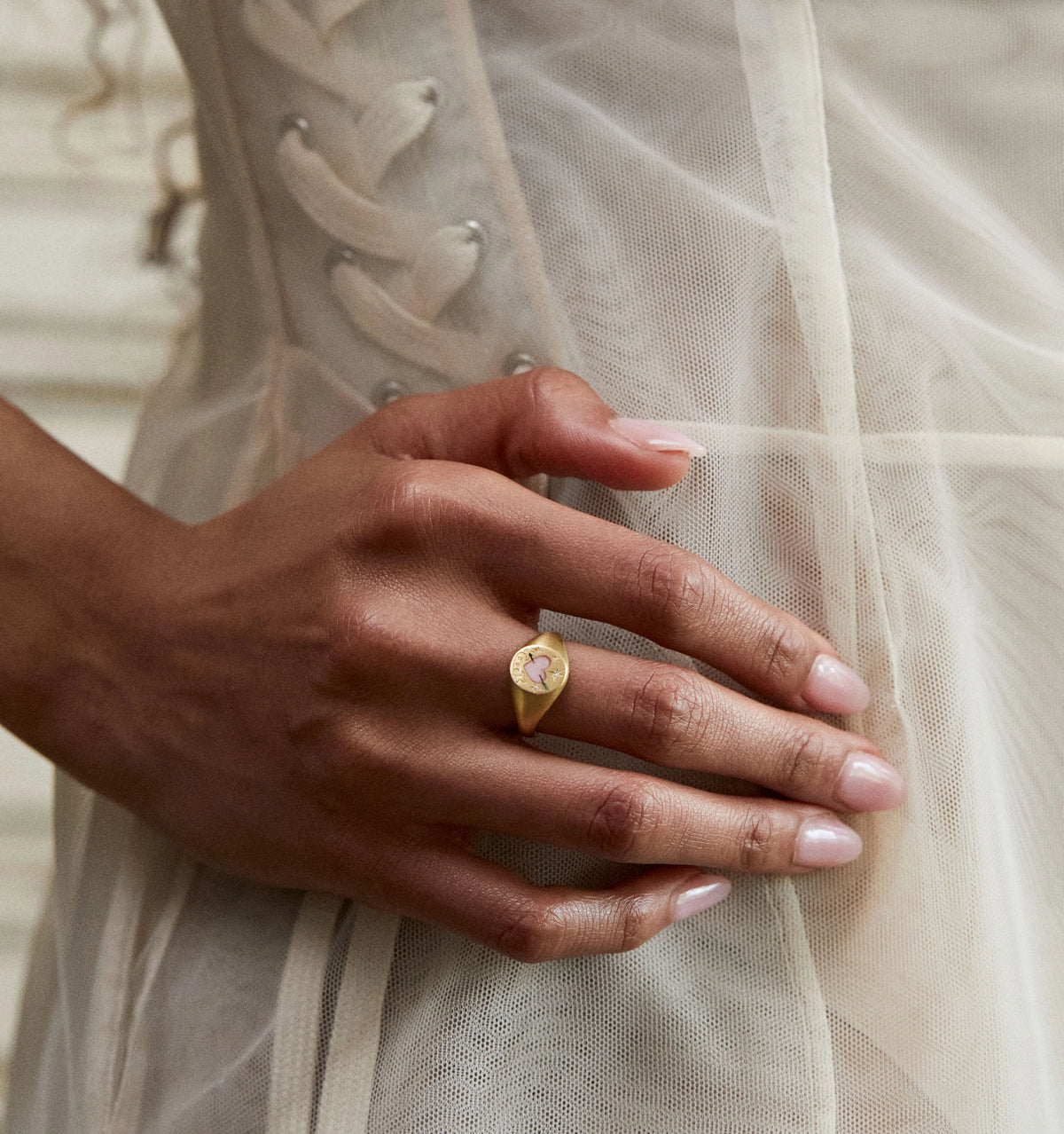 Woman wearing a gold ring featuring a floral design on her finger, with a sheer fabric background.