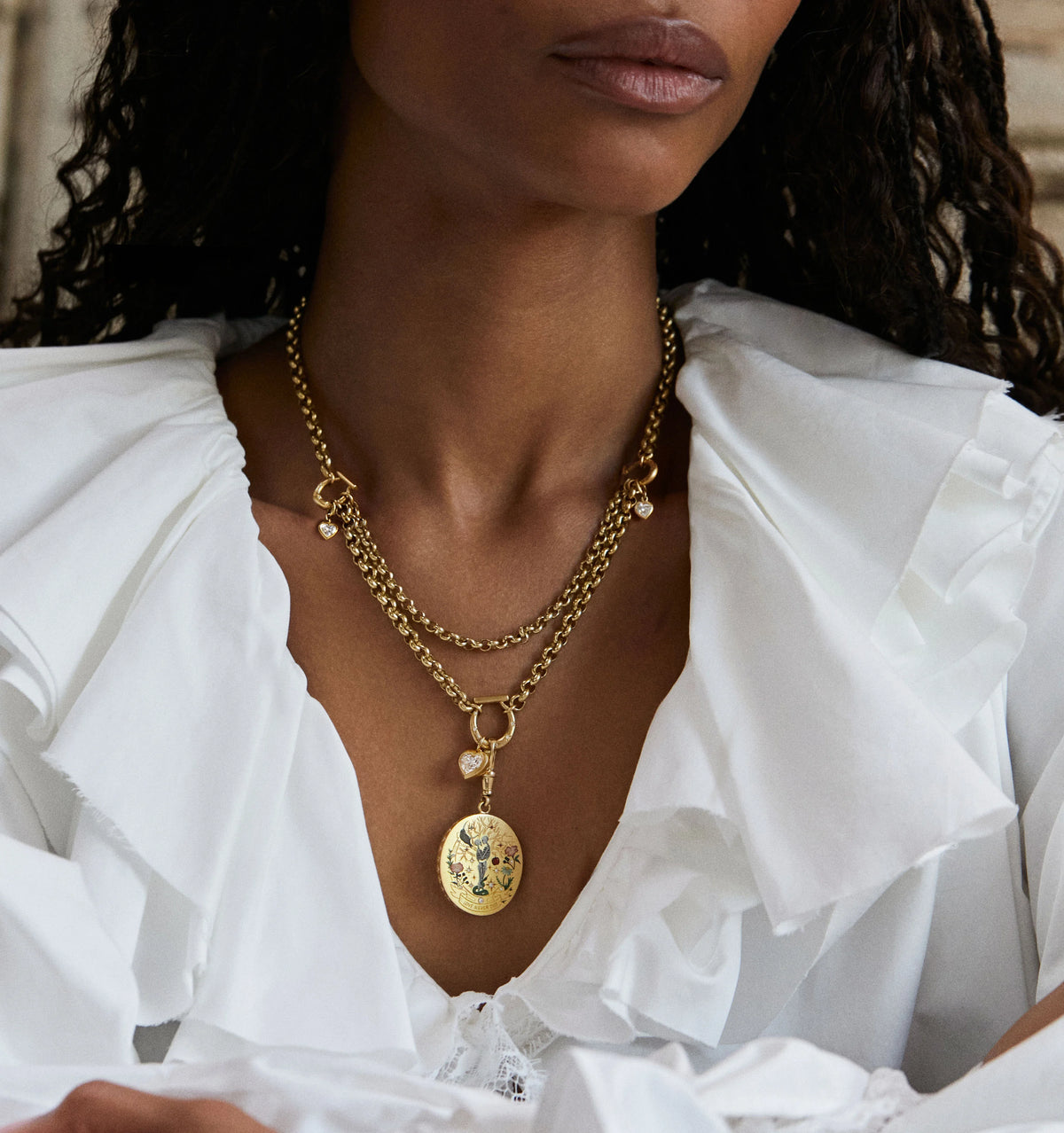 Woman wearing a gold layered necklace with an engraved pendant and a small charm, styled with a white ruffled top.
