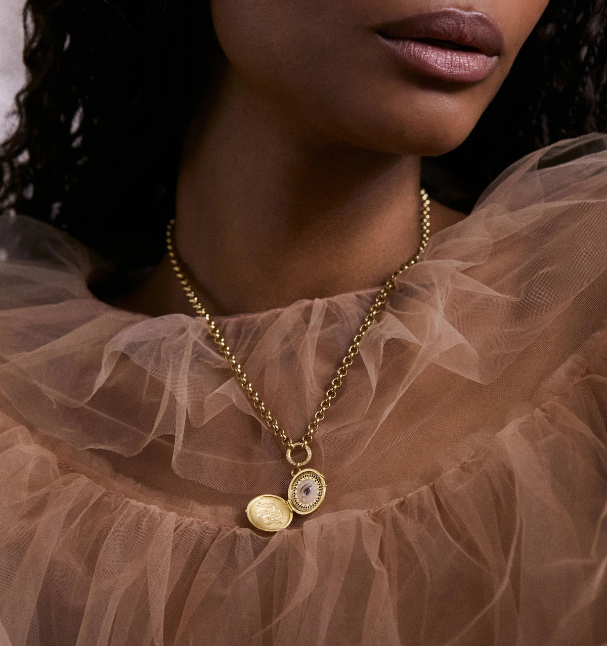 Woman wearing a gold chain necklace featuring an eye motif and an engraved charm, styled with a sheer, ruffled top.