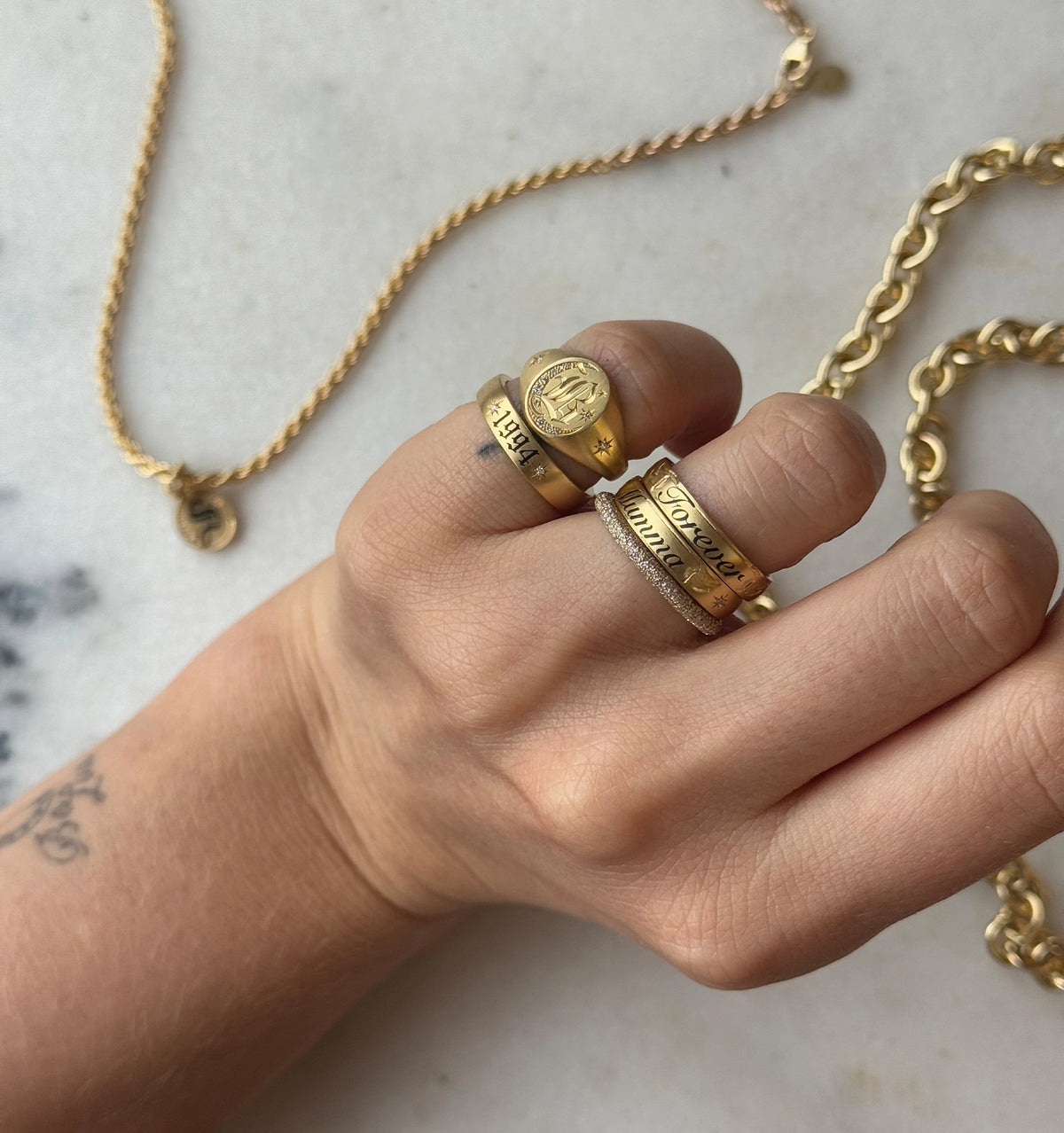 Woman wearing multiple gold rings, including engraved and textured designs, with a gold chain necklace in the background.