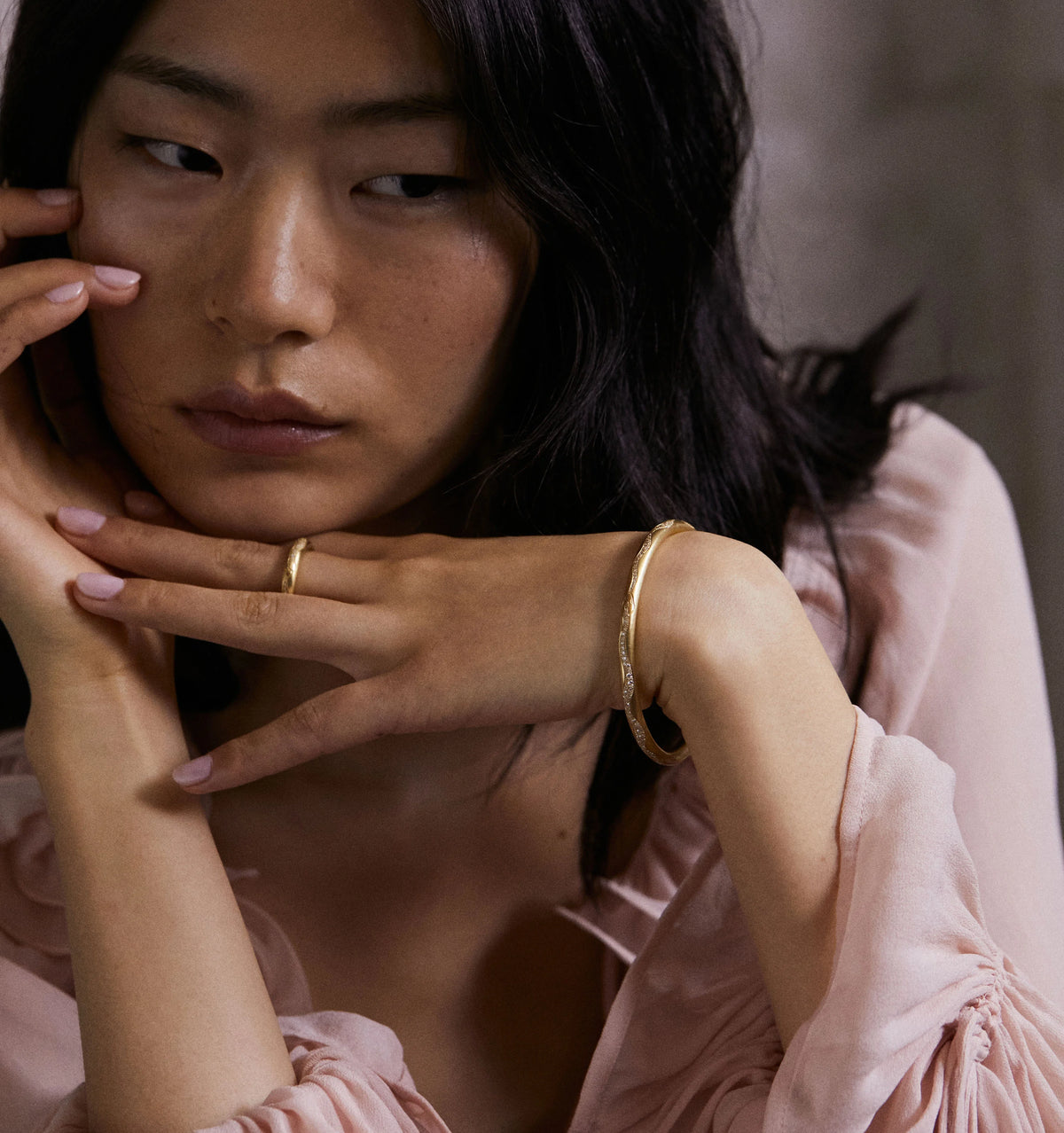 Woman wearing a gold bracelet and ring, resting her chin on her hand in a soft-lit setting.