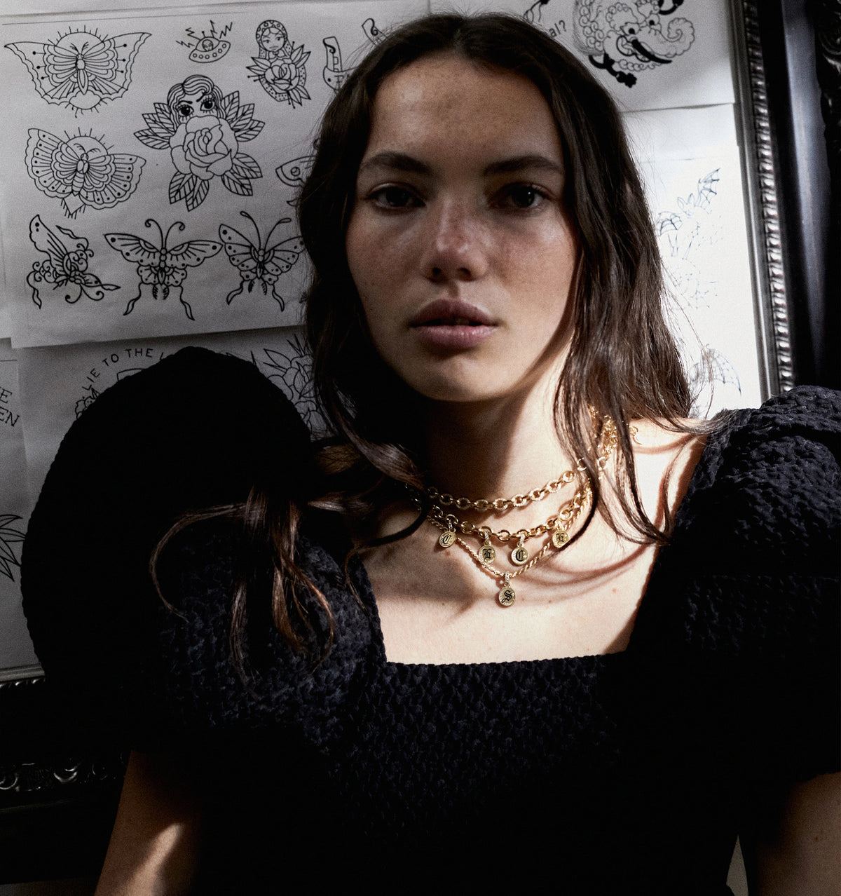 Woman wearing layered gold necklaces against a backdrop of tattoo designs.
