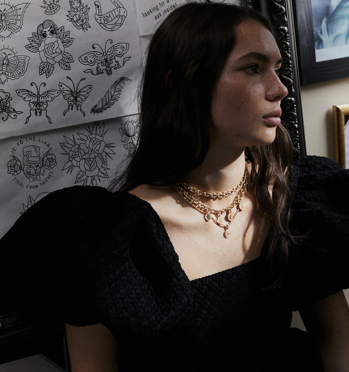 Woman wearing layered gold necklaces against a backdrop of tattoo sketches and framed art.