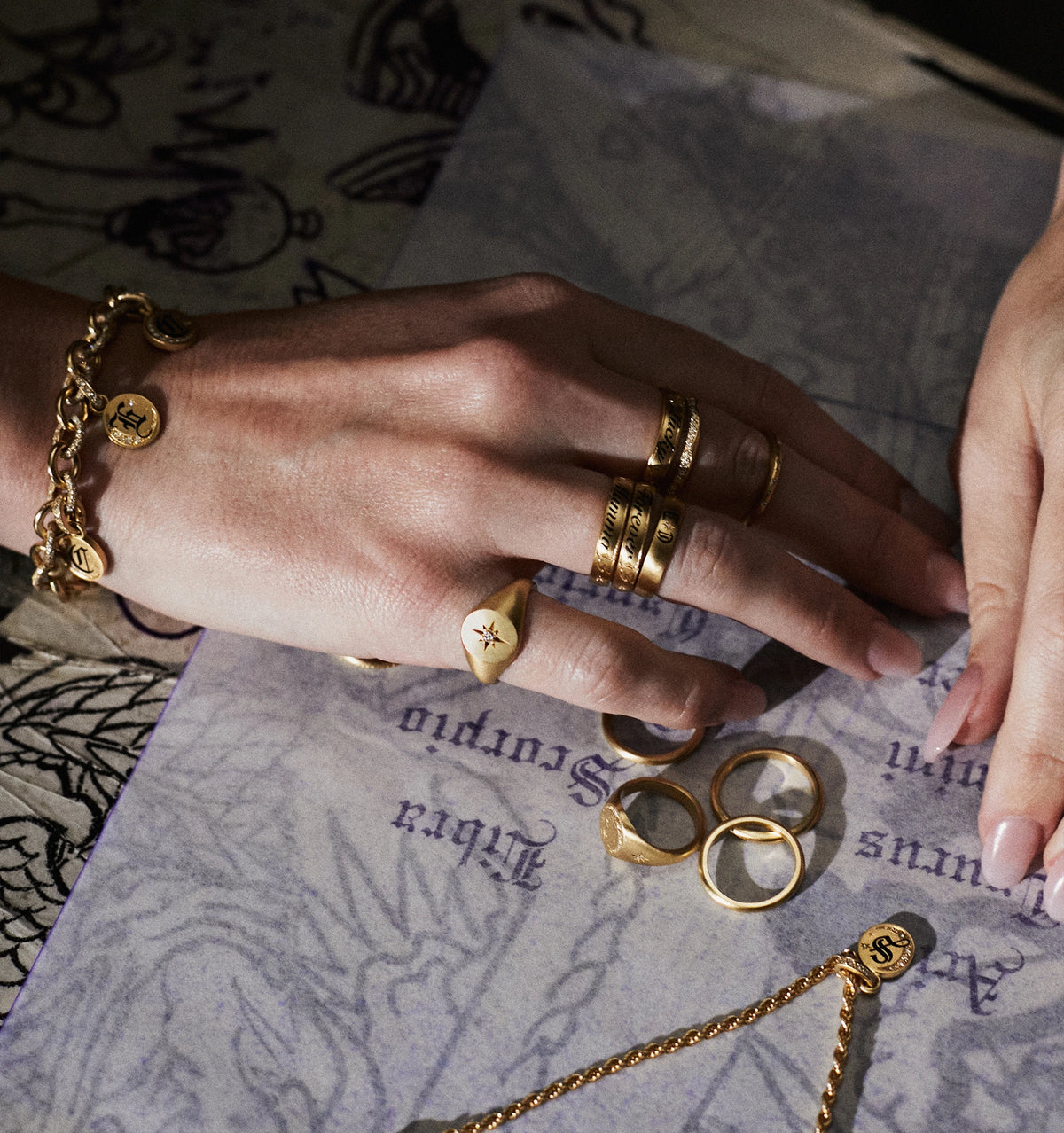 Woman wearing a gold bracelet and multiple gold rings on her hand, with a gold pendant necklace resting on a textured surface.