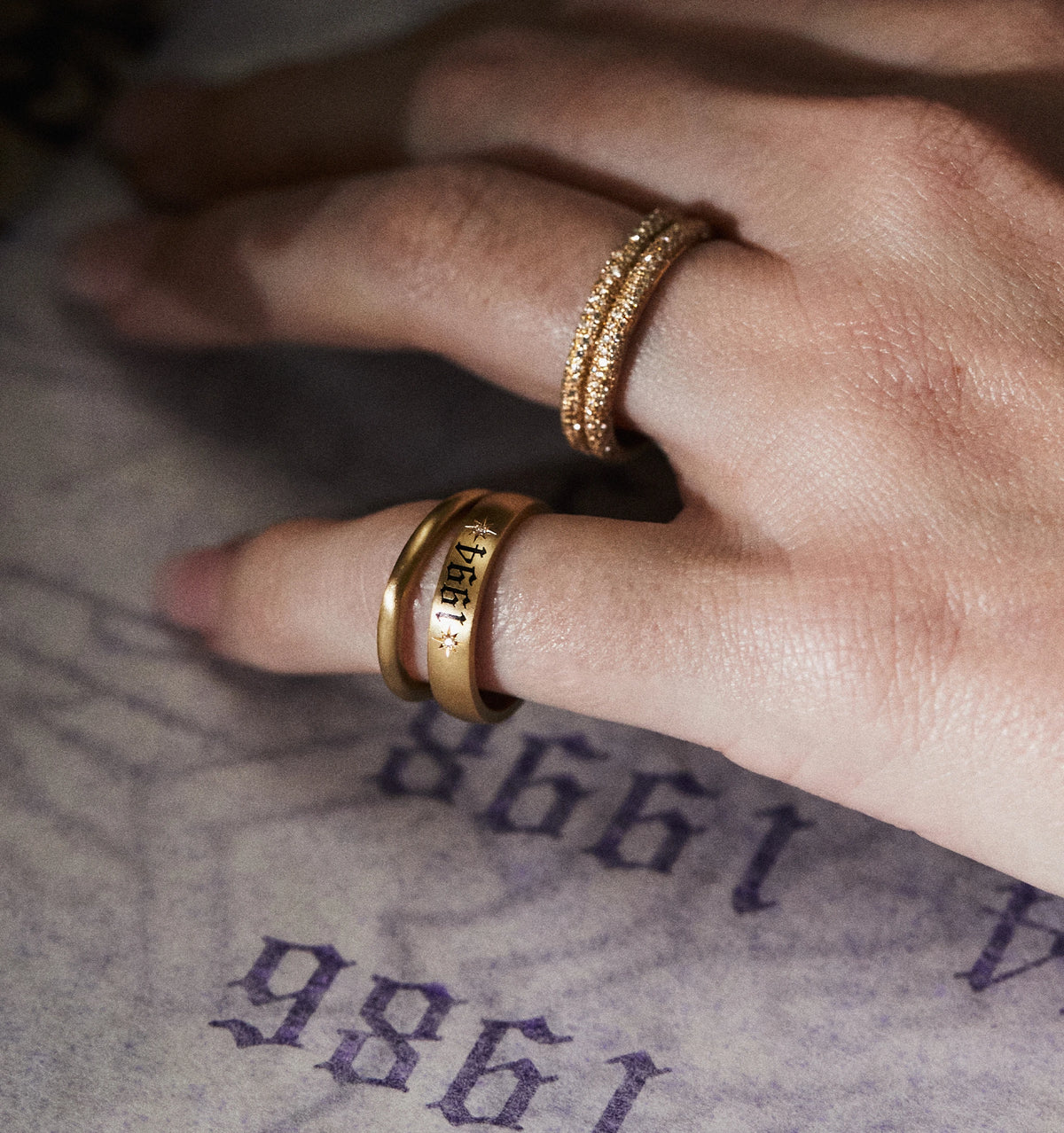 Woman wearing two gold rings, one engraved with "Bali" and the other a braided design, resting on a textured surface.