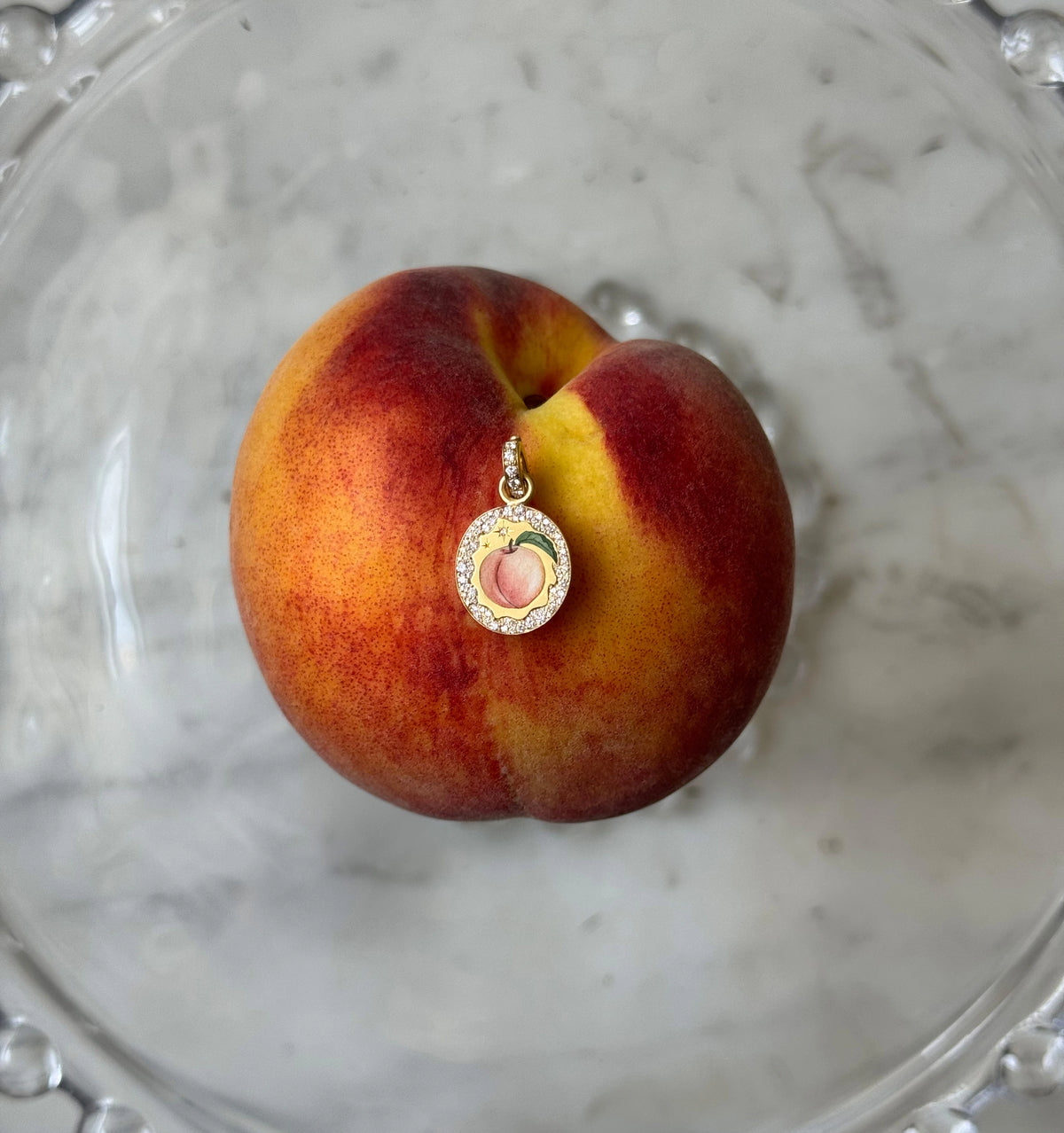 Gold pendant featuring a hand-painted peach design with a diamond-encrusted border, resting on a fresh peach.