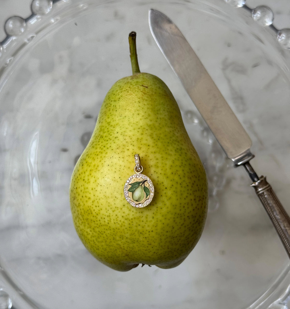 Gold pendant featuring a pear in enamel, displayed on a green pear in a clear bowl with a knife.