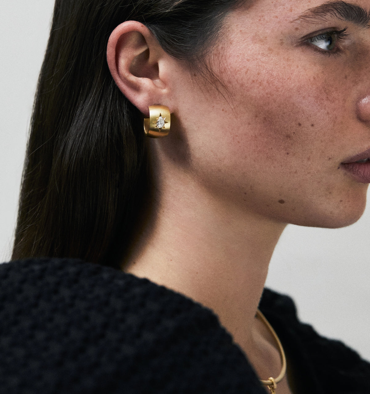 Woman wearing gold hoop earrings featuring a diamond accent, with a visible gold necklace.
