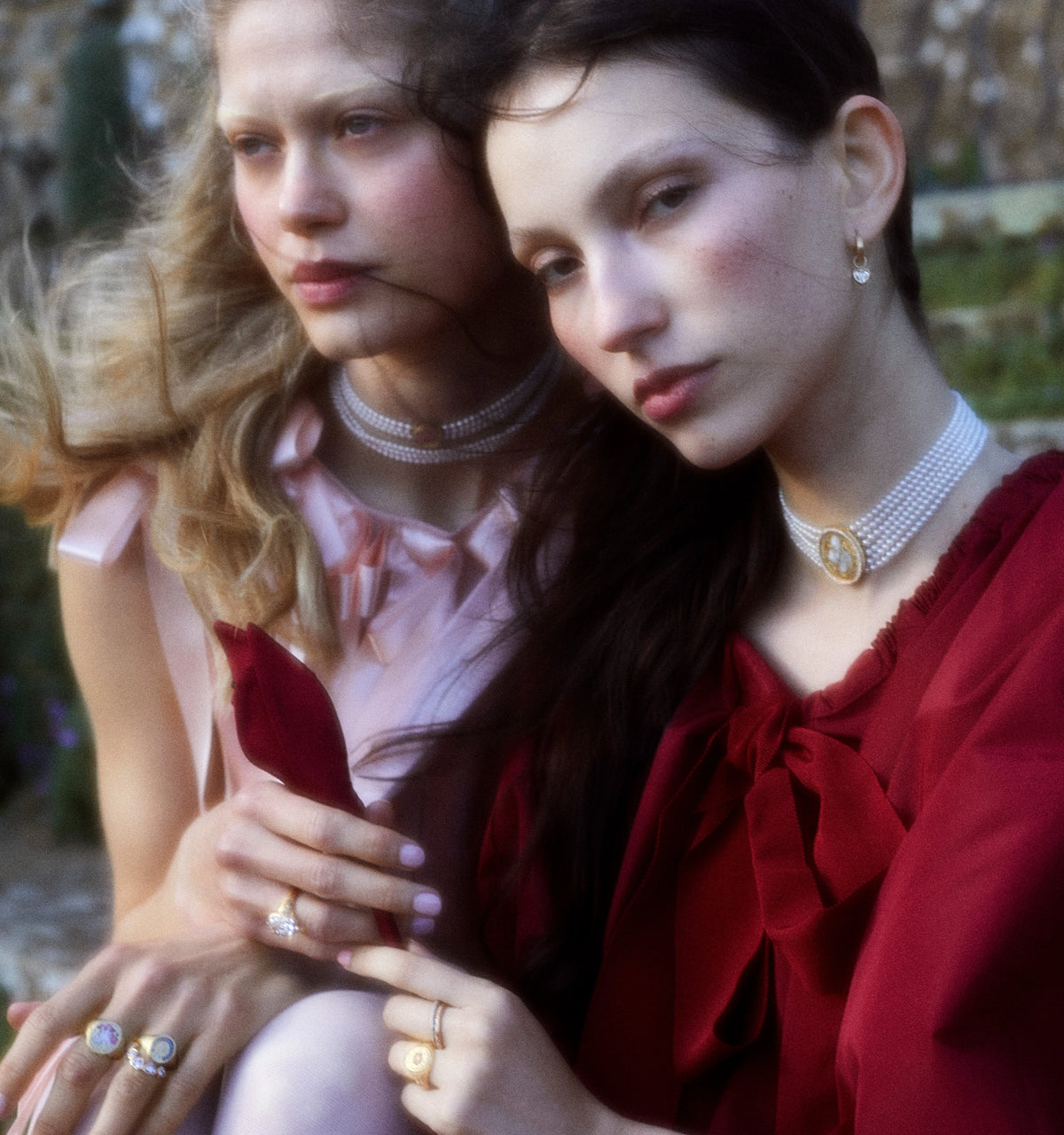 Woman wearing a gold choker and gold ring, with another woman wearing a gold pendant and multiple gold rings, outdoors.