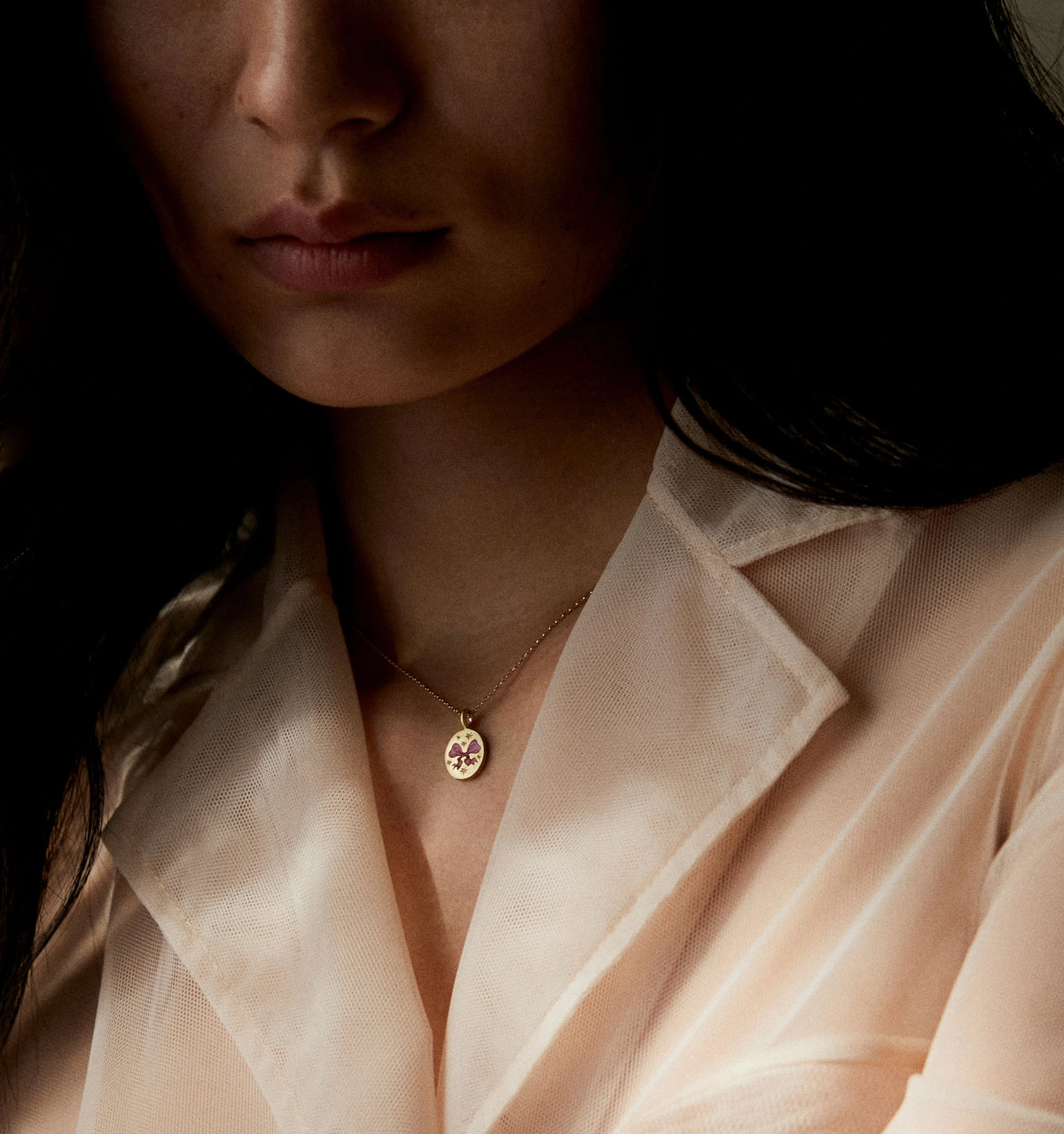 Woman wearing a gold pendant with an enamel bow design against a soft beige blouse.