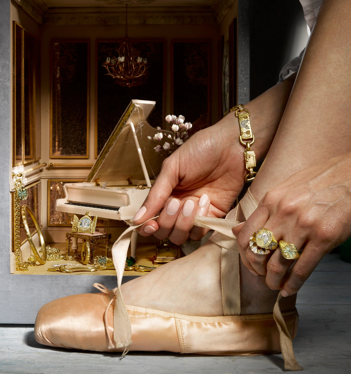 Woman wearing a gold bracelet and two gold rings while tying ballet shoes, with a grand piano in the background.