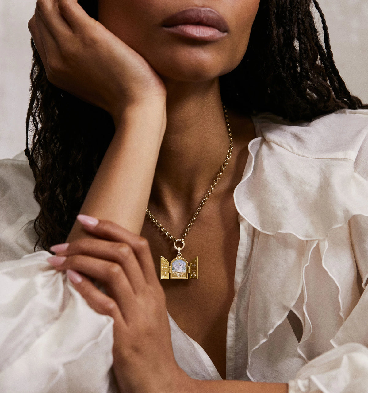 Woman wearing a gold necklace with a pendant featuring a decorative door design, posing with her hand on her chin.