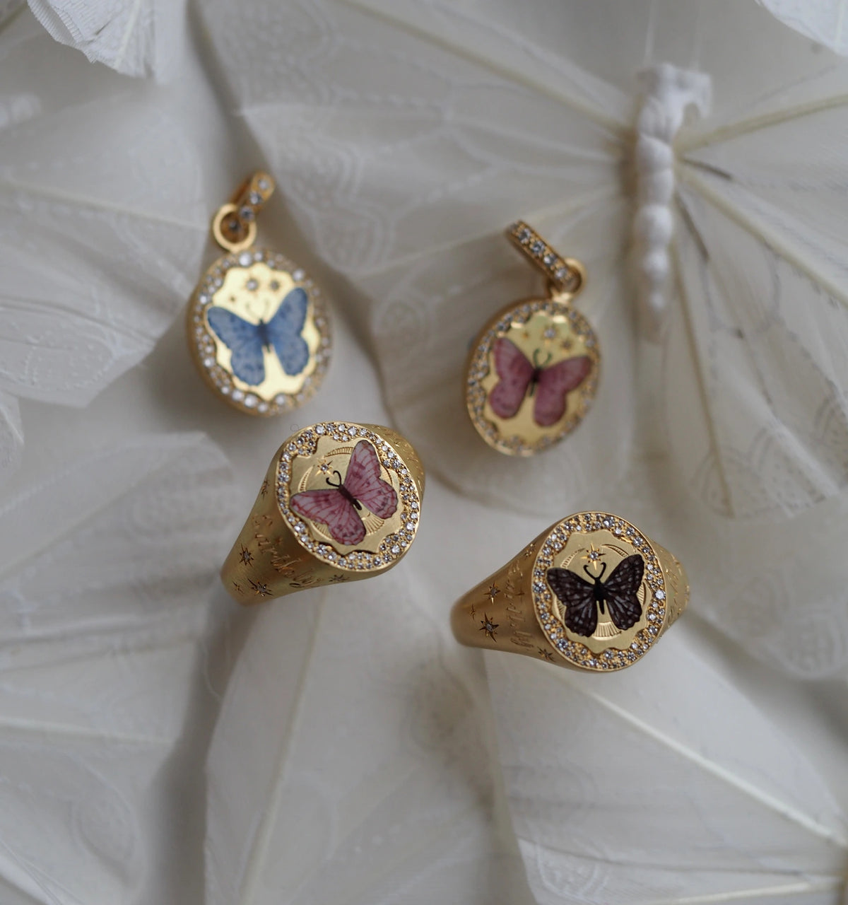 Gold rings and charms featuring butterfly motifs in enamel, set against a delicate fabric background.