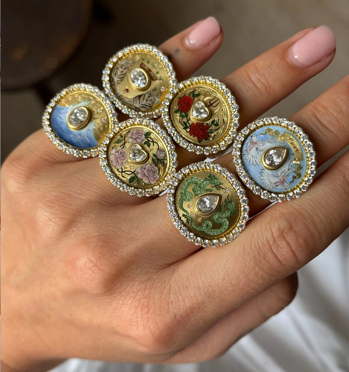 Woman wearing five gold rings, each featuring colourful enamel designs and surrounded by diamonds.