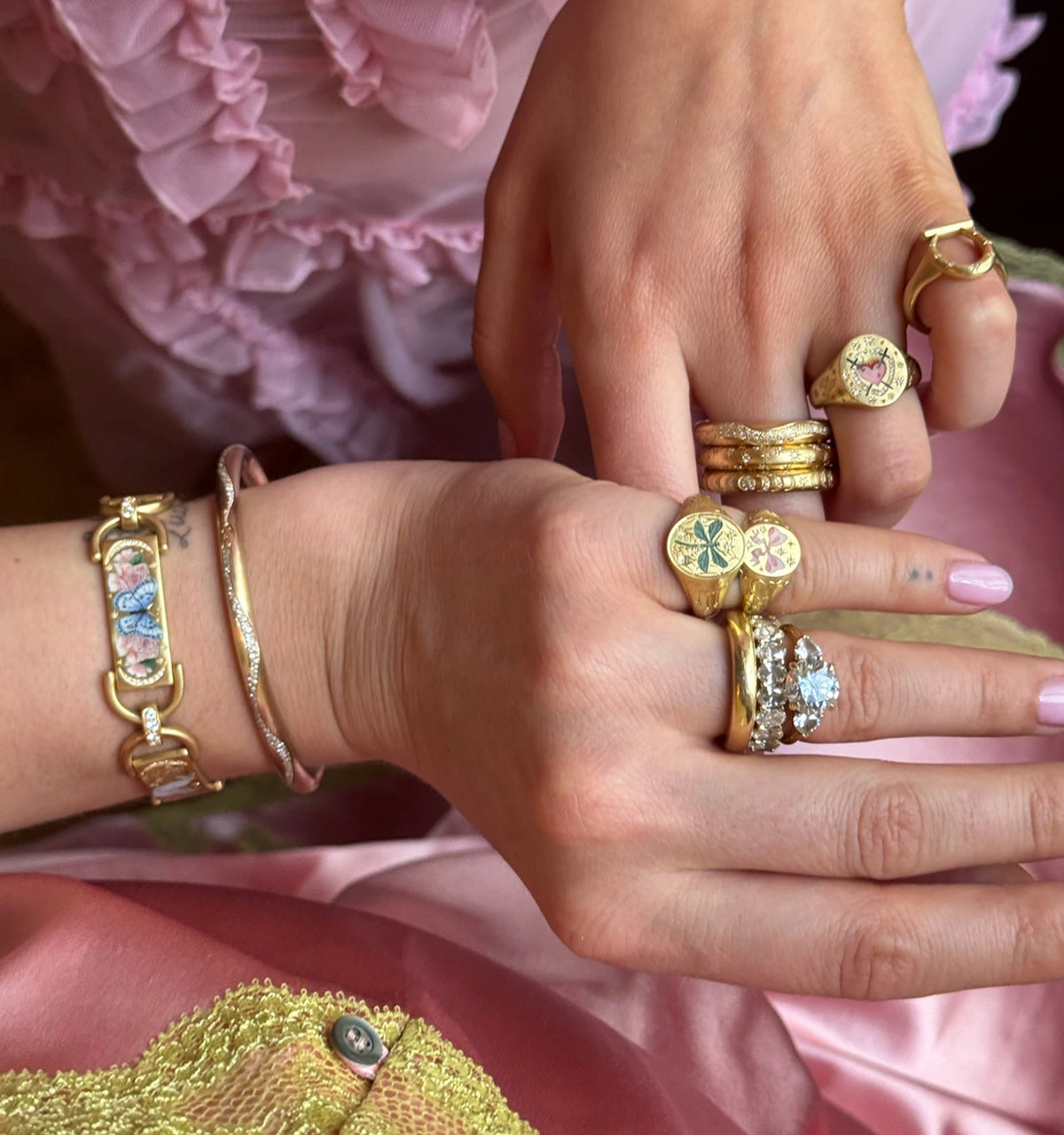 Woman wearing multiple gold rings, including one with pink and green enamel motifs, and a gold bracelet with floral enamel details.