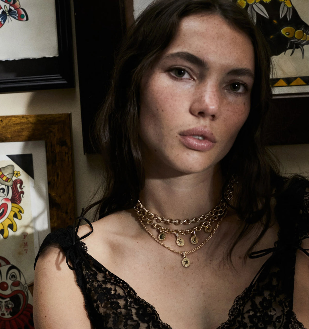 Woman wearing layered gold chains with pendants against a backdrop of framed artwork.