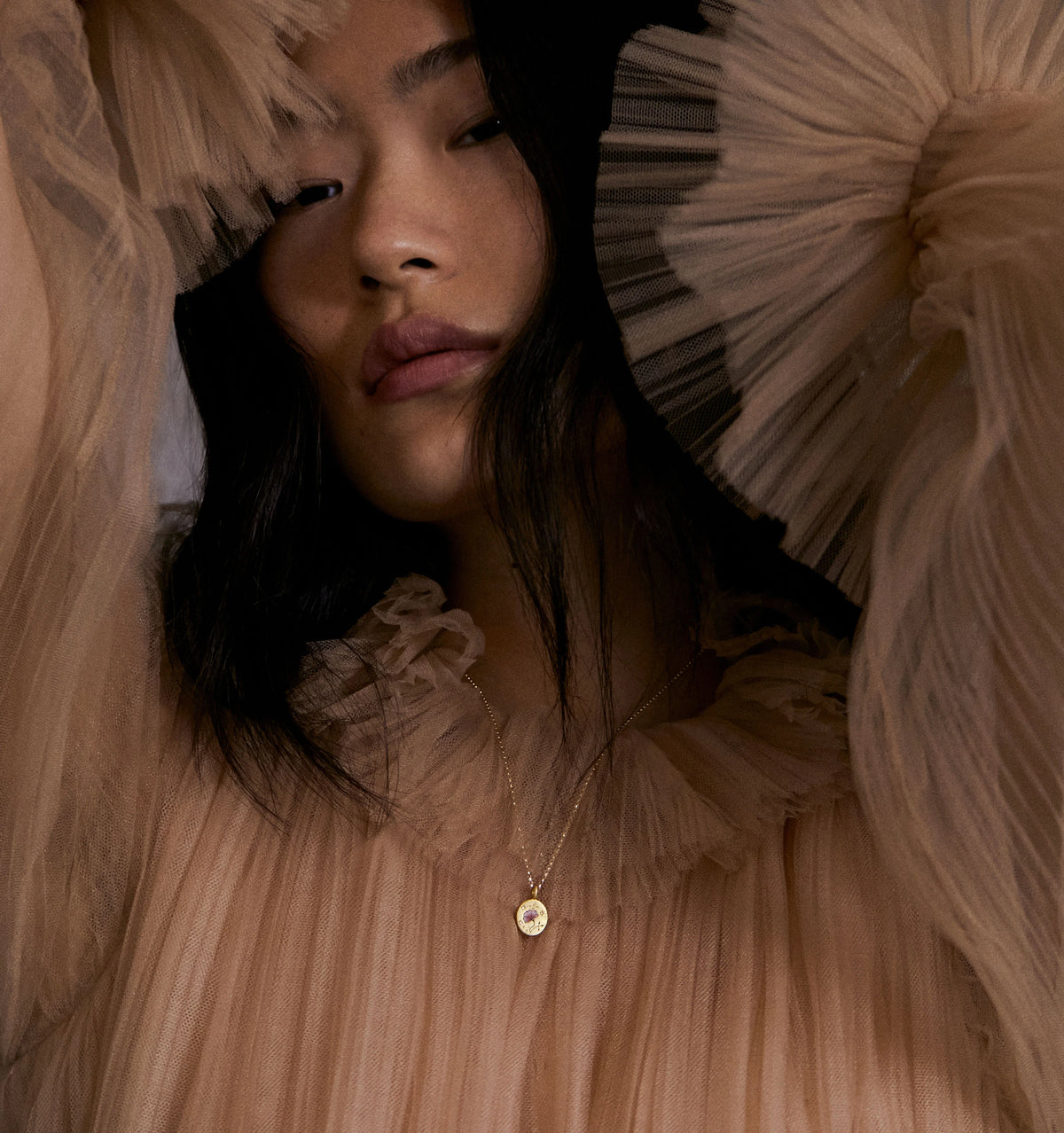 Woman wearing a gold pendant necklace with a circular charm on a delicate chain, styled with a ruffled nude blouse.