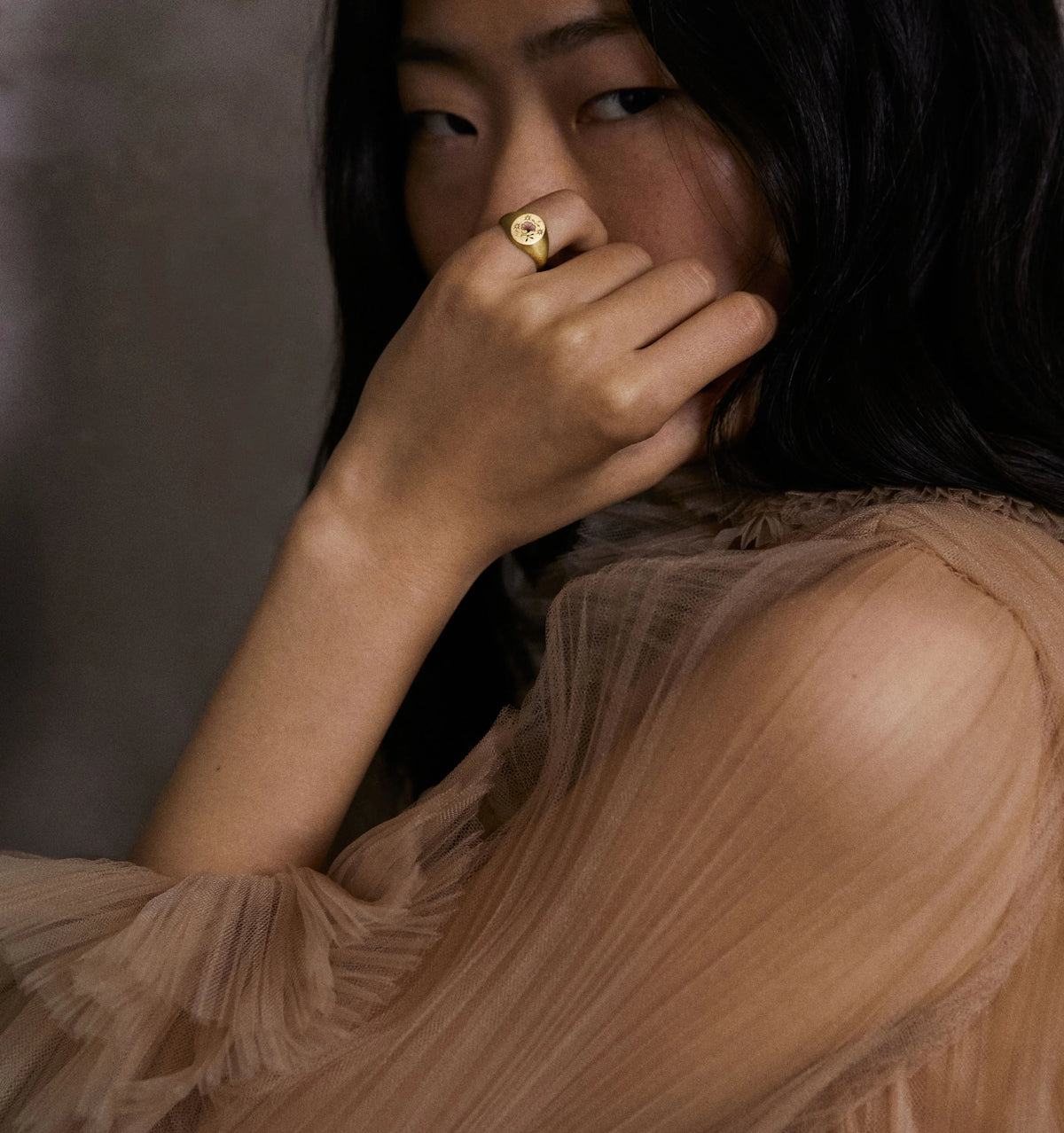 Woman wearing a gold ring while partially covering her face with her hand, dressed in a sheer, beige top.
