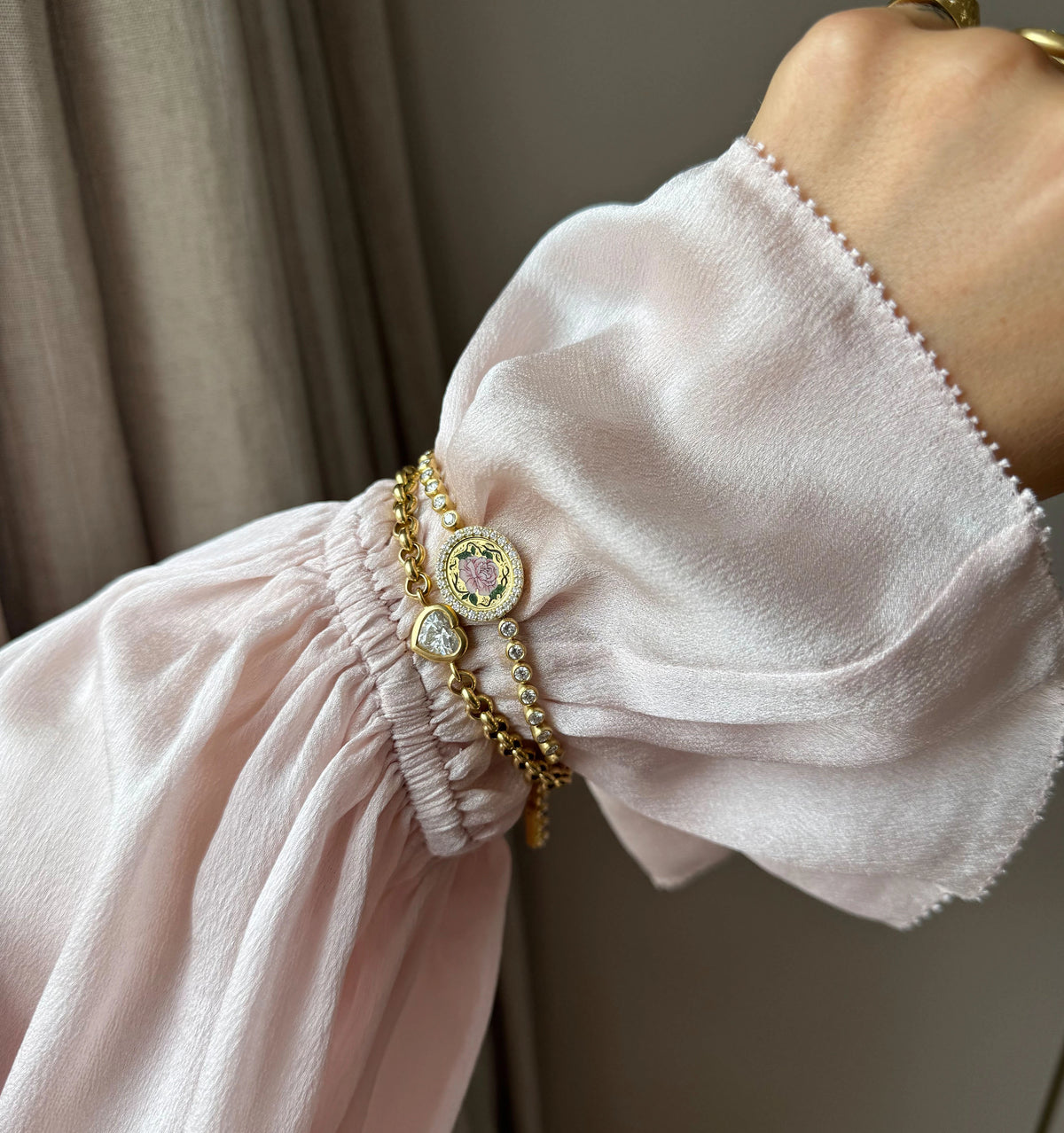 Woman wearing multiple gold bracelets, including a charm bracelet and a beaded design, with a soft pink blouse.