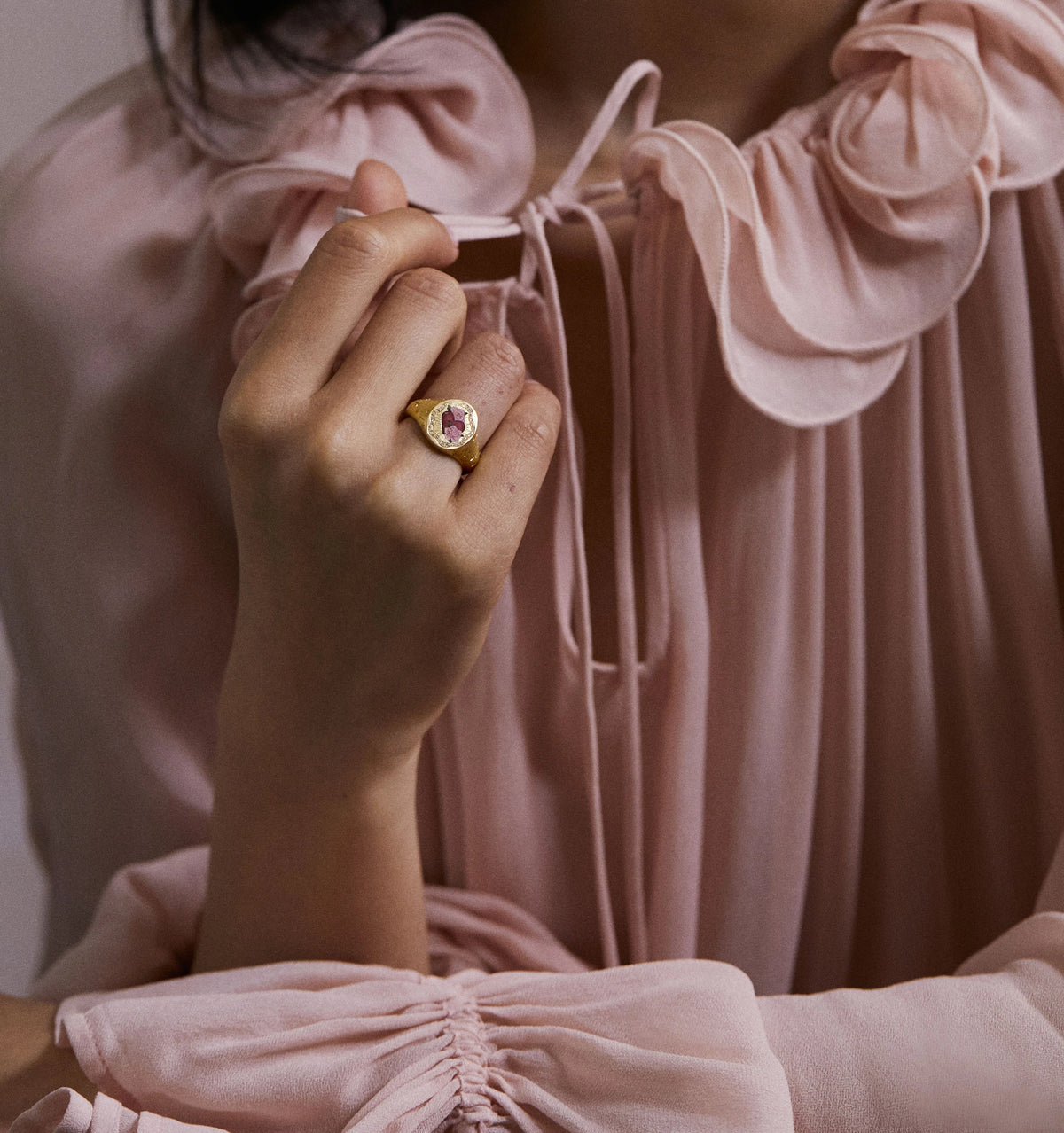Woman wearing a gold ring featuring a red rose, holding her hand near a ruffled pink blouse.