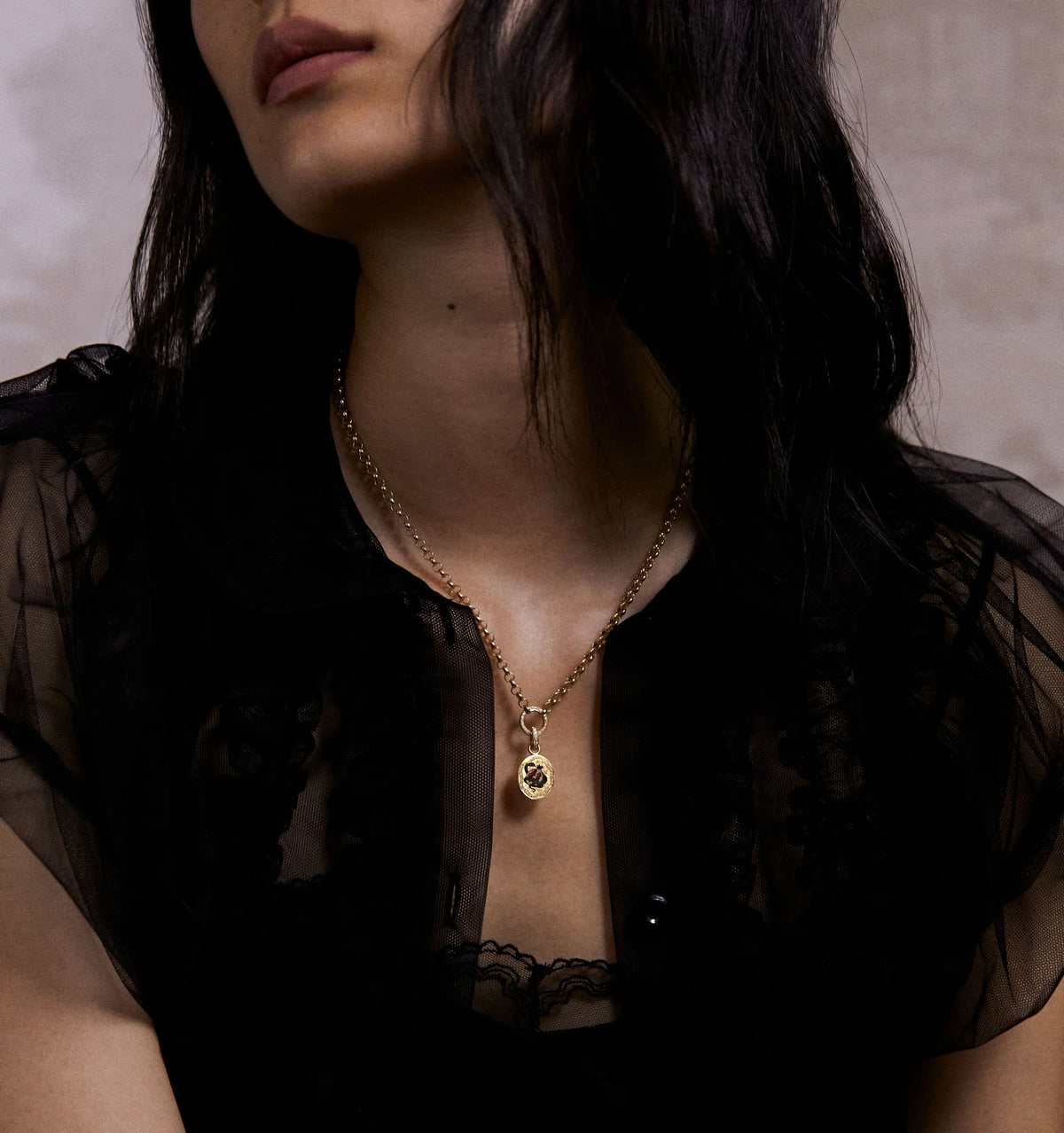 Woman wearing a gold necklace with a pendant featuring red enamel and a sheer black blouse.