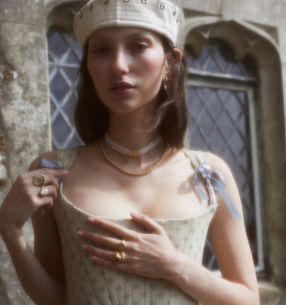 Woman wearing a gold chain necklace and multiple gold rings, with a light blue dress and a cream-coloured hat.