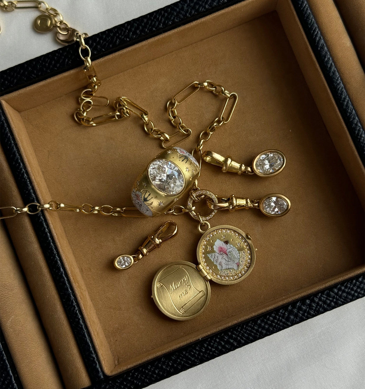 Gold jewellery set including an engraved locket, a ring with two diamonds, and decorative gold links, arranged on a textured surface.