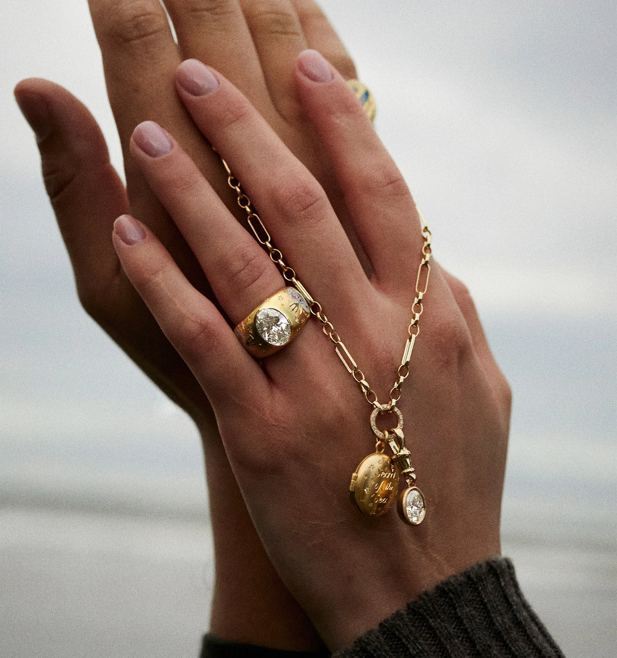 Woman wearing a gold ring and gold chain necklace with a charm, hands intertwined against a cloudy background.