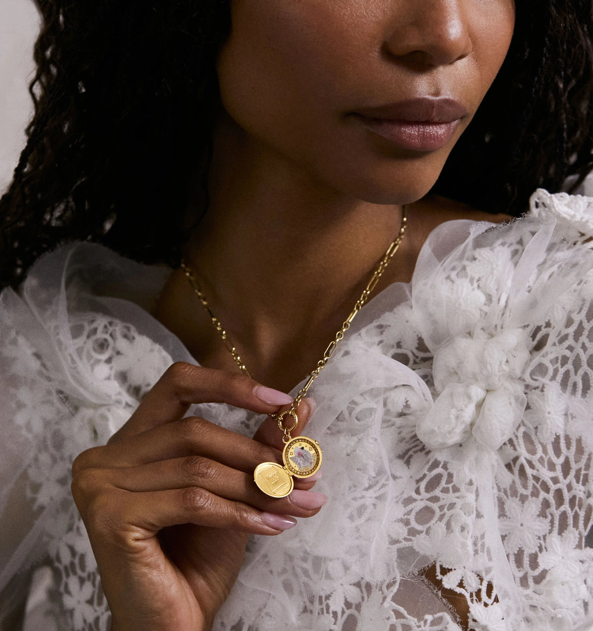 Woman wearing a gold necklace with a circular pendant featuring a clear stone, holding it near her face.