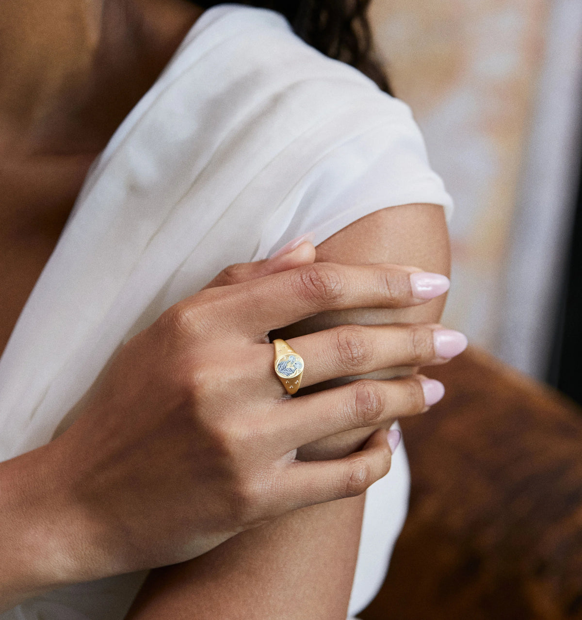 Woman wearing a gold ring featuring a blue stone, resting her hand on her arm while dressed in a white top.