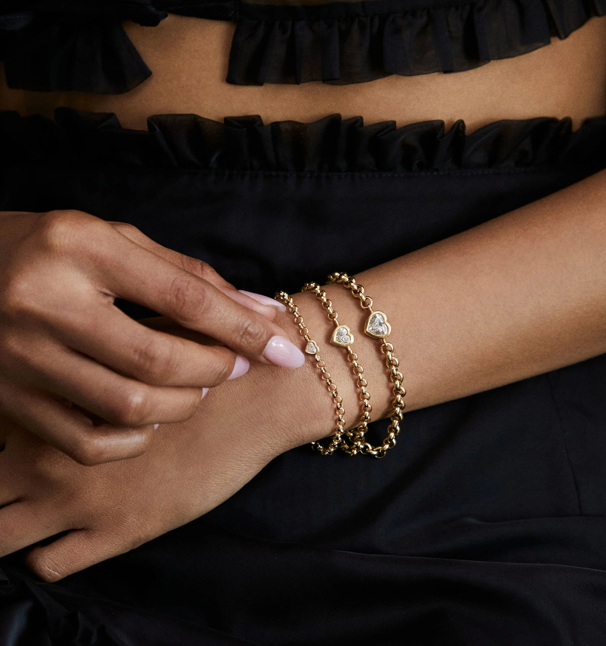 Woman wearing three gold bracelets featuring heart-shaped pendants on a black background.