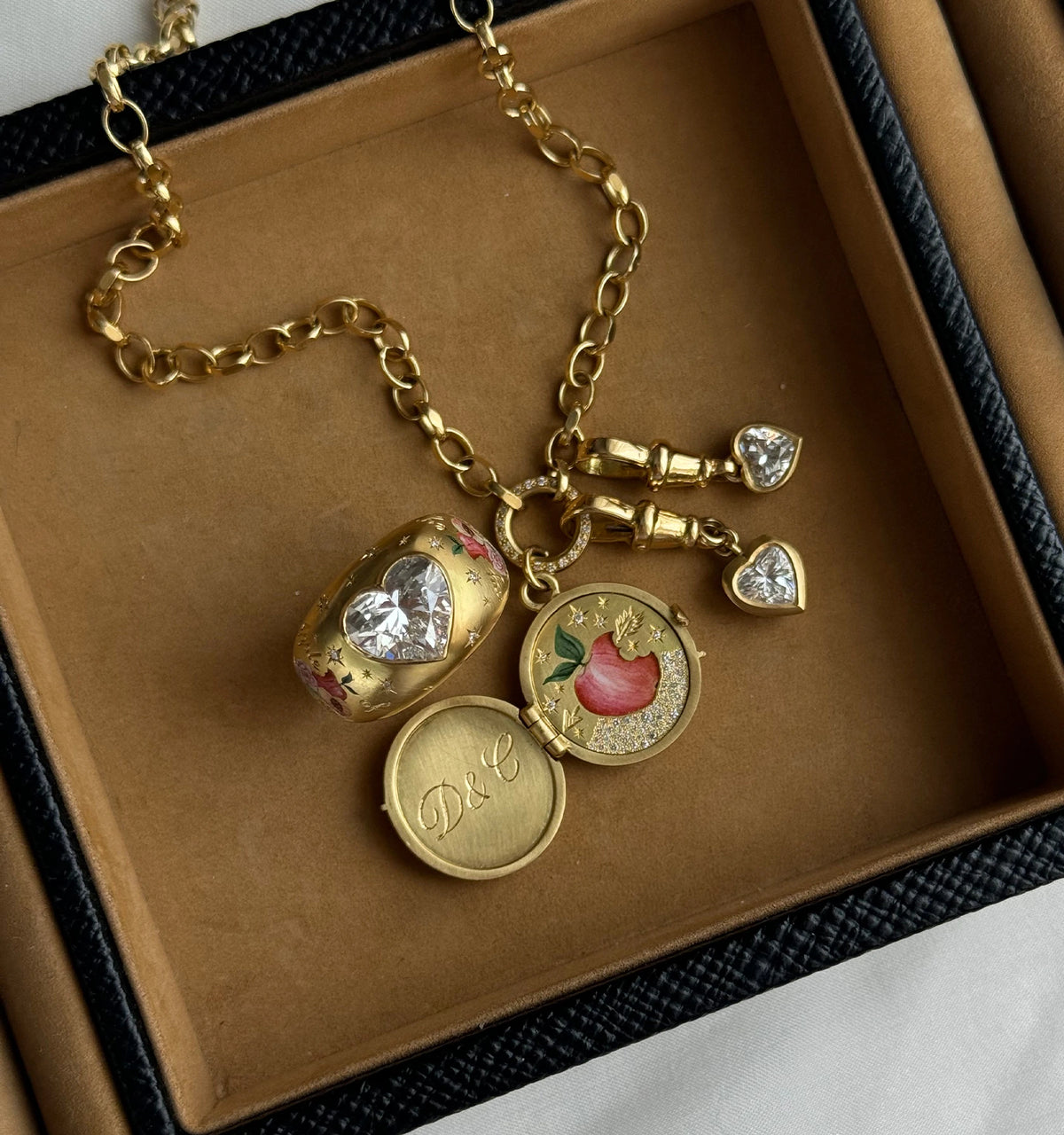 Gold necklace featuring a heart-shaped charm with a clear stone, an engraved locket, and decorative enamel detailing.