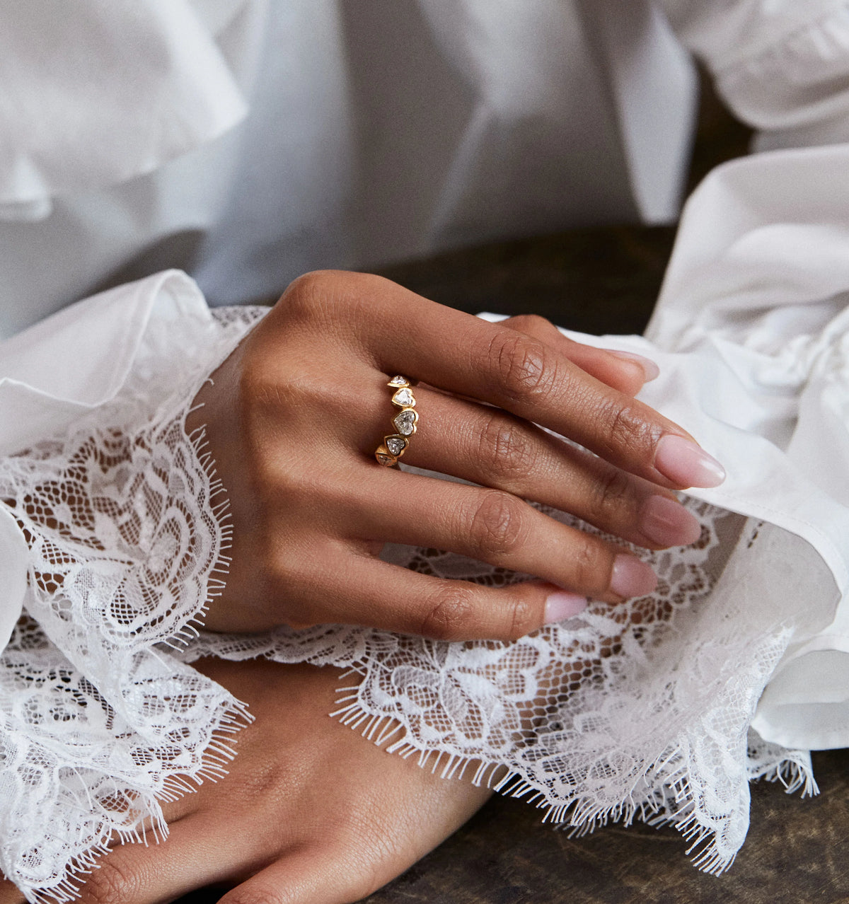 Woman wearing a gold ring with heart-shaped designs and diamonds, resting on a lace-covered surface.