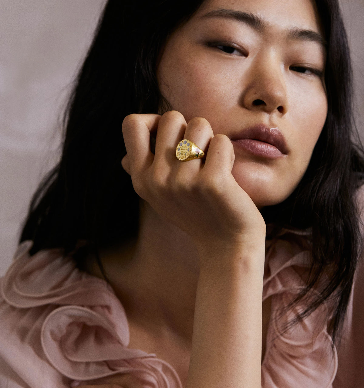 Woman wearing a gold ring with engraved patterns, resting her chin on her hand, against a soft, neutral background.