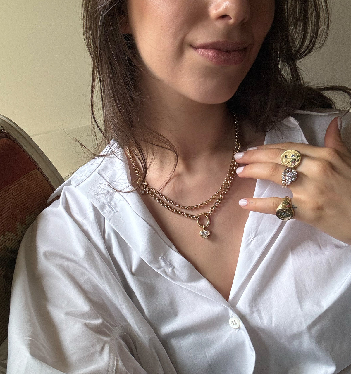 Woman wearing a gold chain necklace with a heart pendant and multiple gold rings, including a gold coin ring and a gem-encrusted ring.