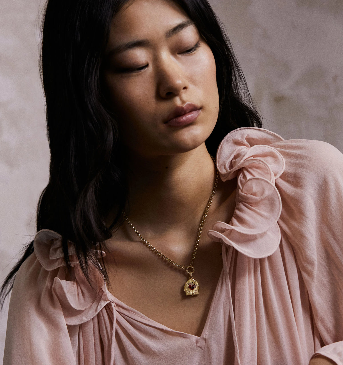 Woman wearing a gold necklace with a charm featuring a red stone, styled with a pink ruffled blouse.