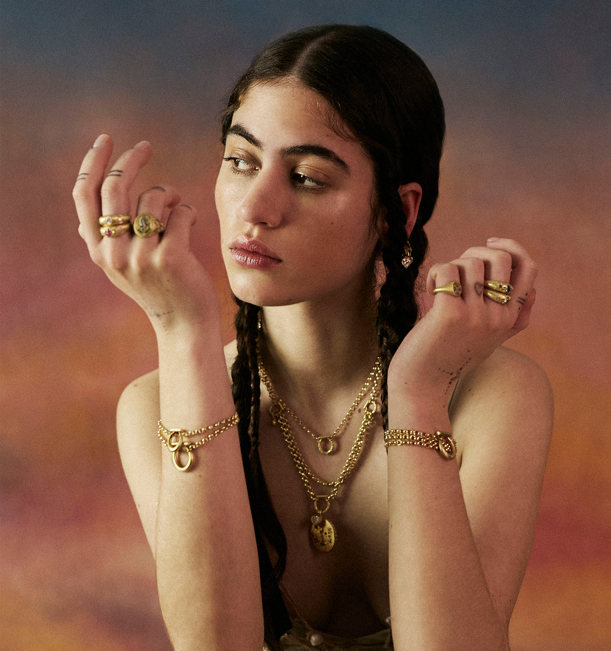 Woman wearing multiple gold rings and gold layered necklaces, with a warm, textured background.