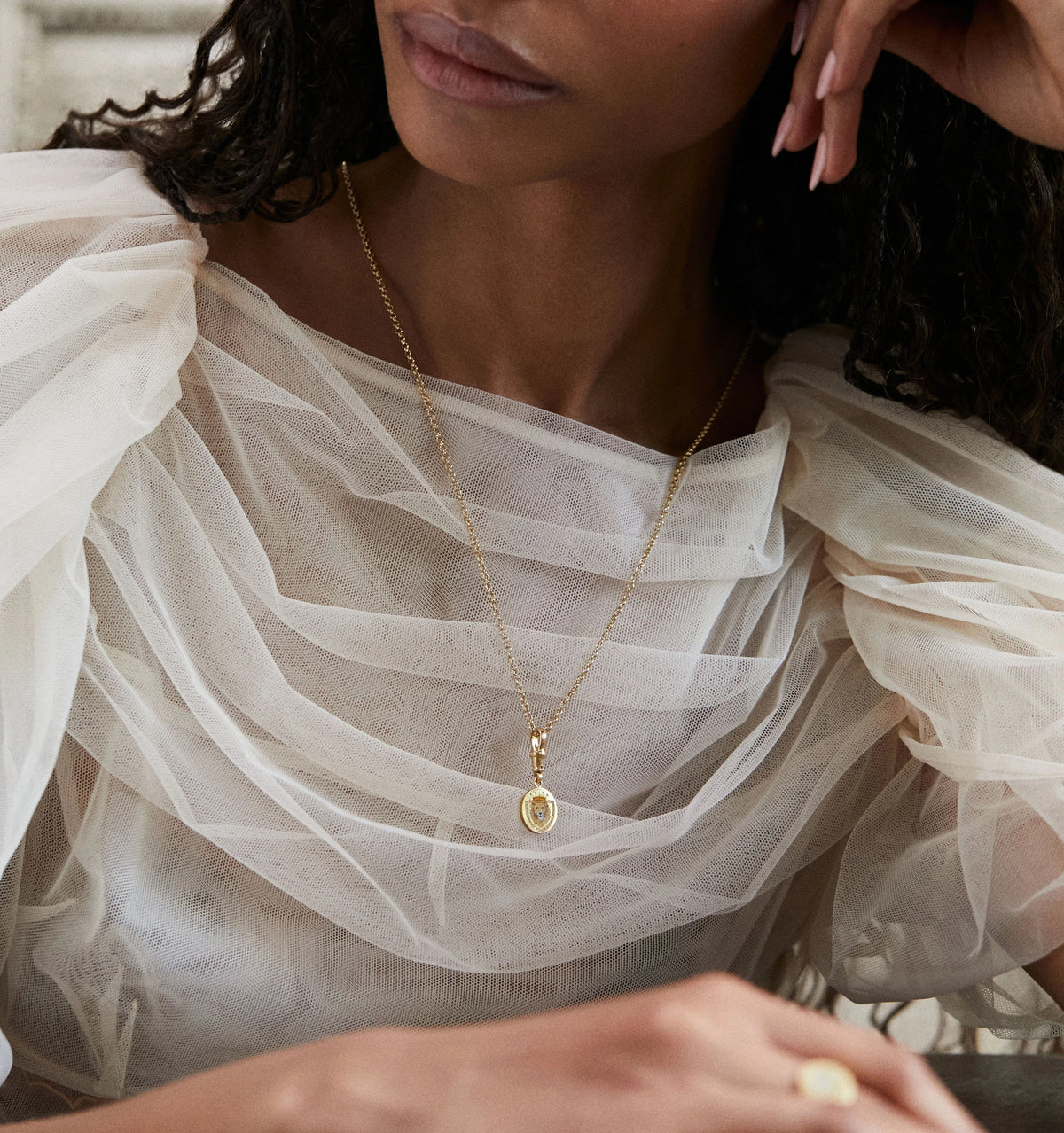 Woman wearing a gold necklace with a pendant on a sheer, white top.