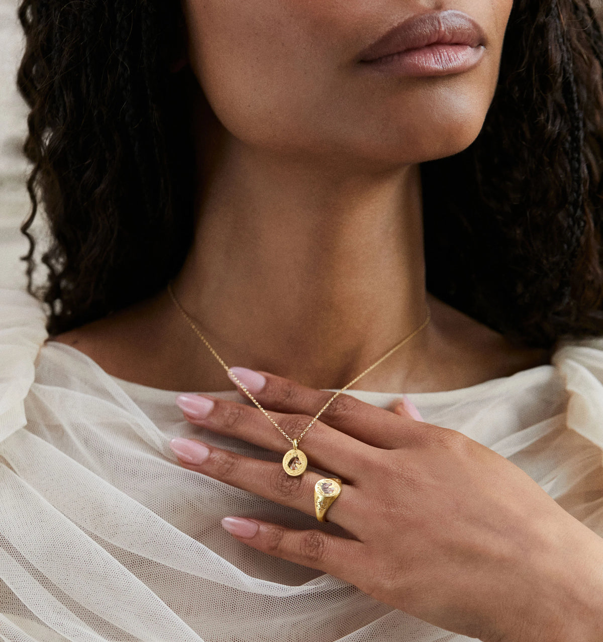 Woman wearing a gold pendant necklace and a gold ring, with soft draped fabric in the background.