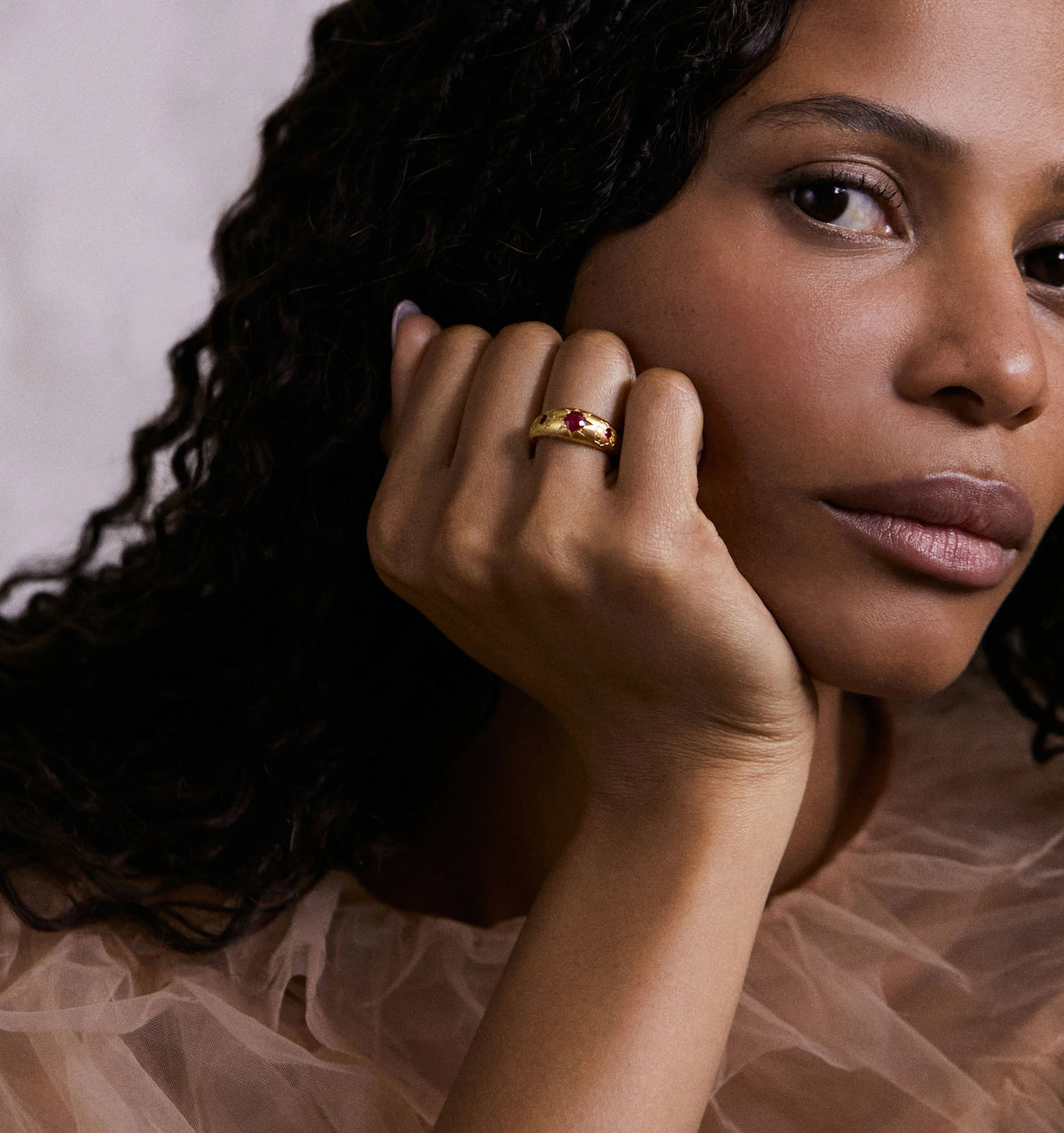 Woman wearing a gold ring featuring rubies, resting her chin on her hand against a soft, neutral background.