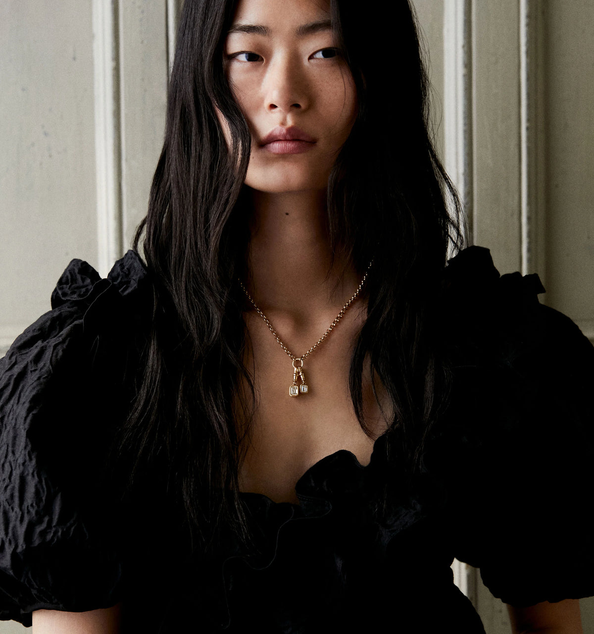 Woman wearing a gold necklace with two lock pendants, dressed in a black puff-sleeved top.