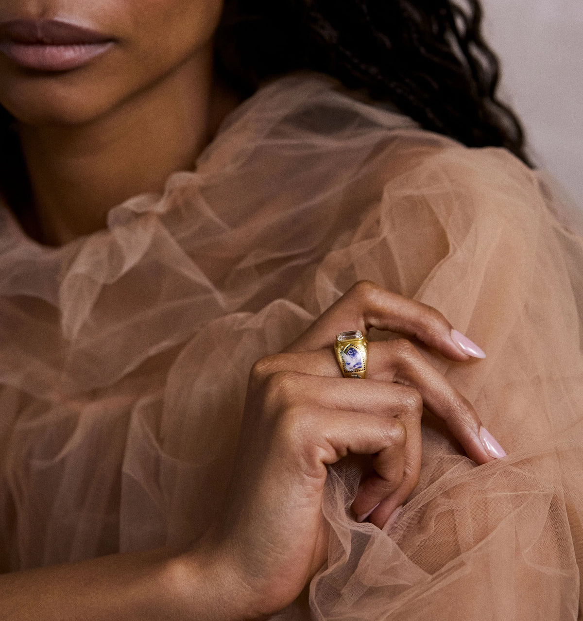 Woman wearing a gold ring featuring blue and white enamel accents, against a soft, sheer fabric background.