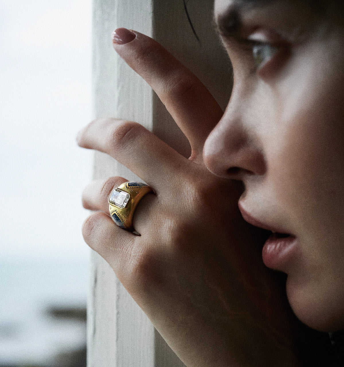 Woman wearing a gold ring with a rectangular stone, gesturing with her hand against a window.