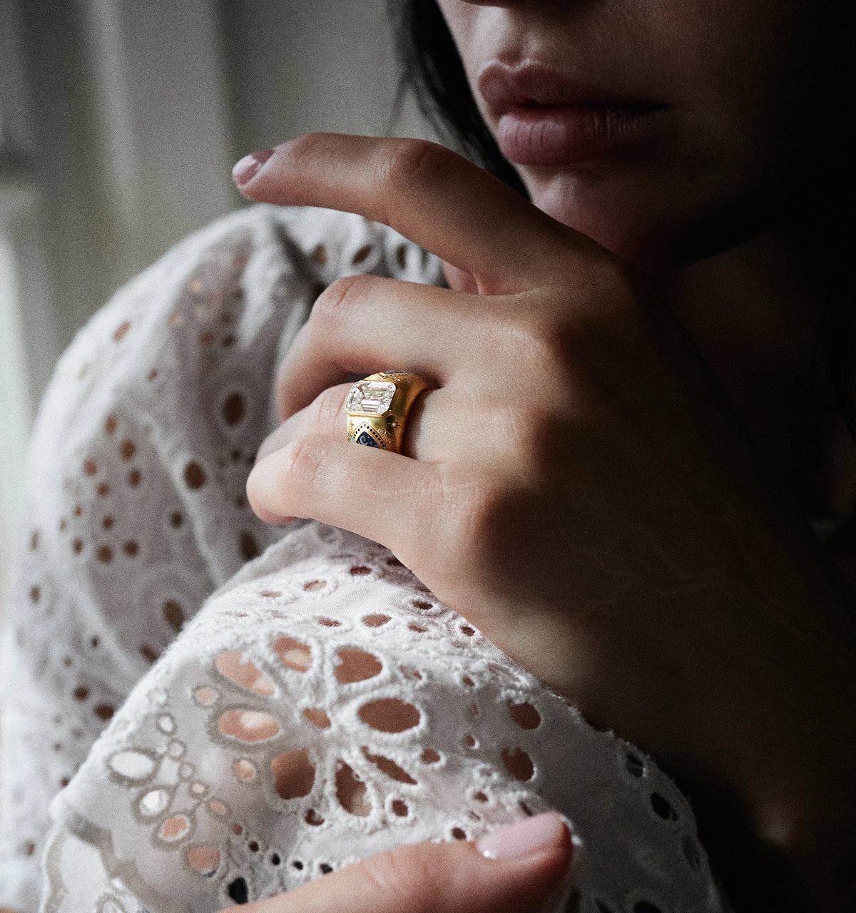 Woman wearing a gold ring set with a rectangular stone and blue accents, resting her hand on a lace top.
