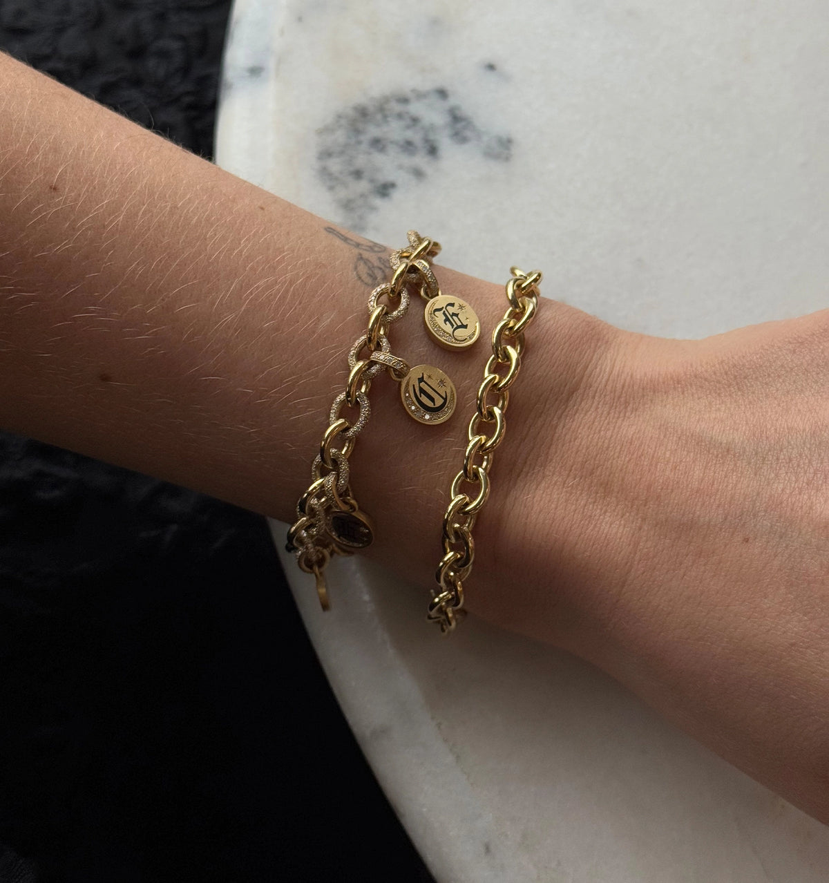 Woman wearing two gold bracelets with circular pendants on a marble surface.