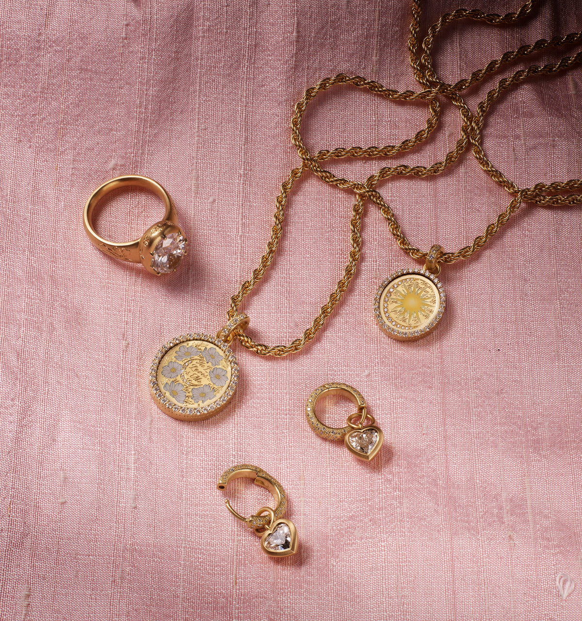 Gold necklace with engraved pendant, gold ring featuring a clear stone, and gold hoop earrings with heart motifs, arranged on pink fabric.