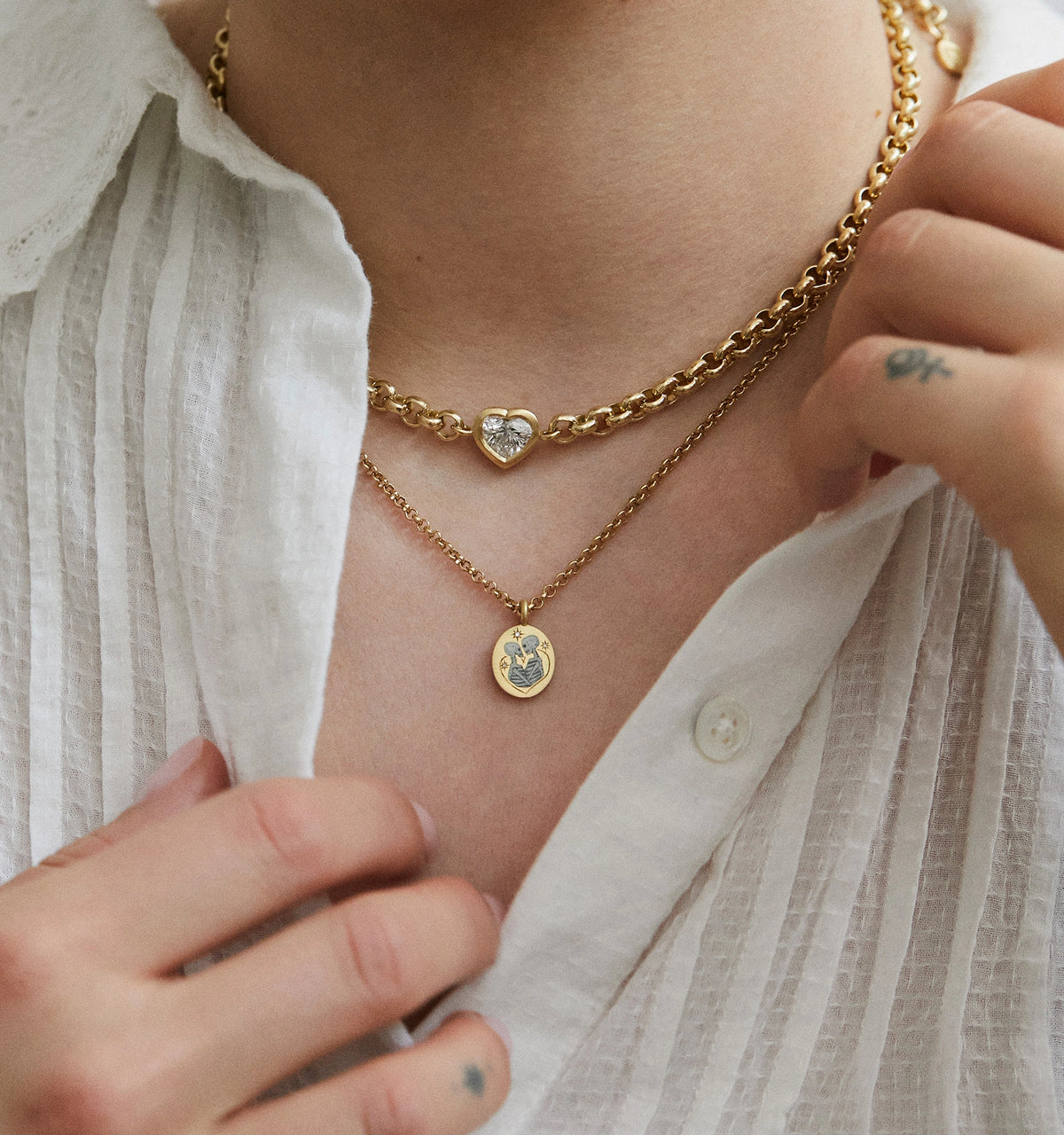 Woman wearing a gold heart-shaped necklace and a gold pendant necklace with an engraved floral design.