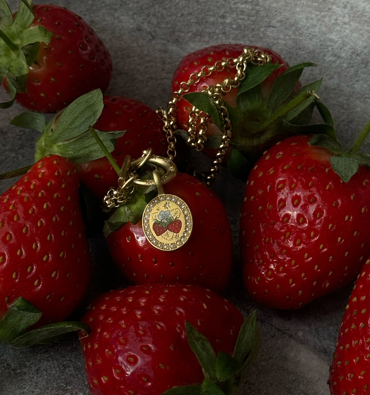 Gold pendant necklace featuring an enamel strawberry design, surrounded by fresh strawberries.